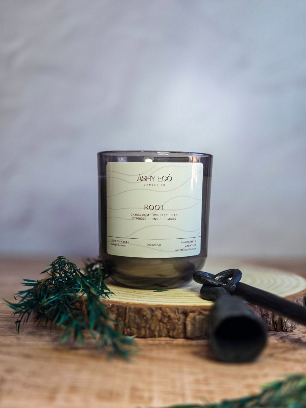 Candle labeled 'Root' by Ashy Ego on a wooden block with greenery and a black tool.