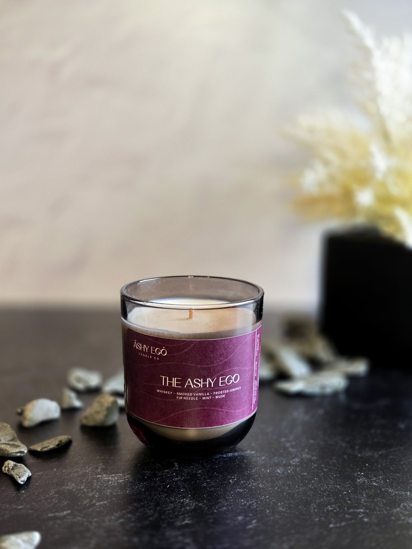 Candle called 'The Ashy Ego' with a purple label on a dark surface, with decorative elements in the background.