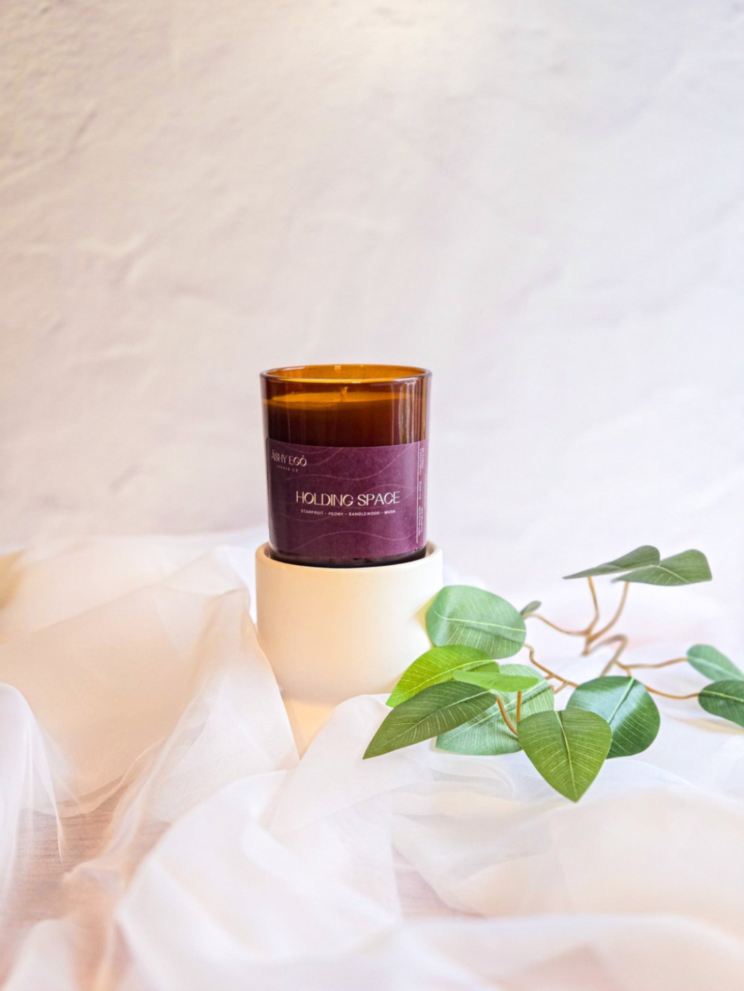 Candle called 'Holding Space' with a purple label on a white surface with green leaves