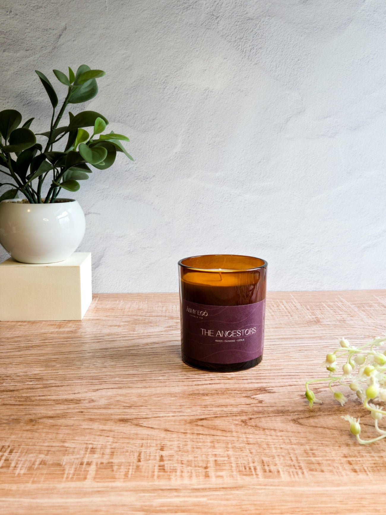 Amber glass candle called 'The Ancestors' with purple label on a wooden surface next to a plant